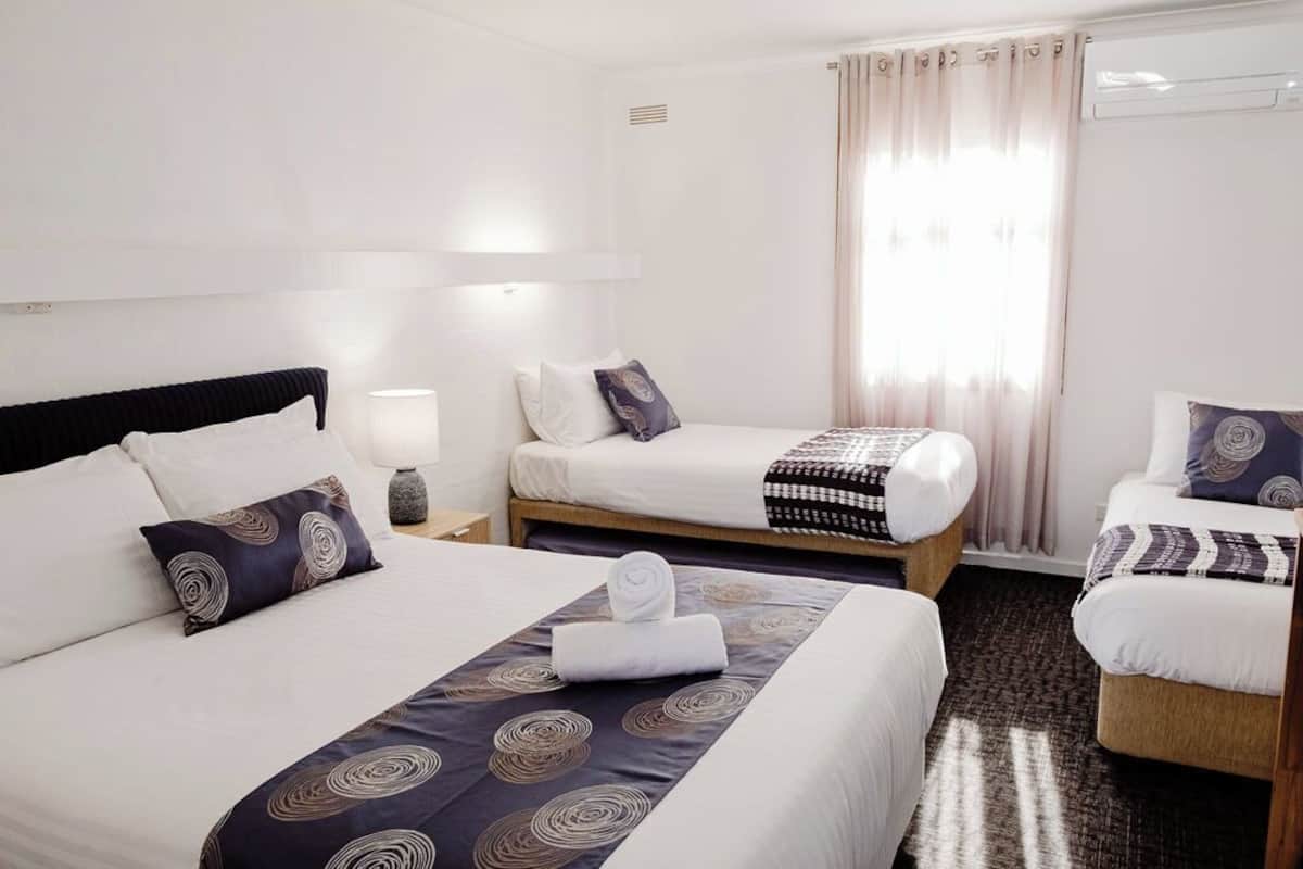 A hotel room with two single beds, a large bed, a bedside table with a lamp, a window with curtains, and a carpeted floor.