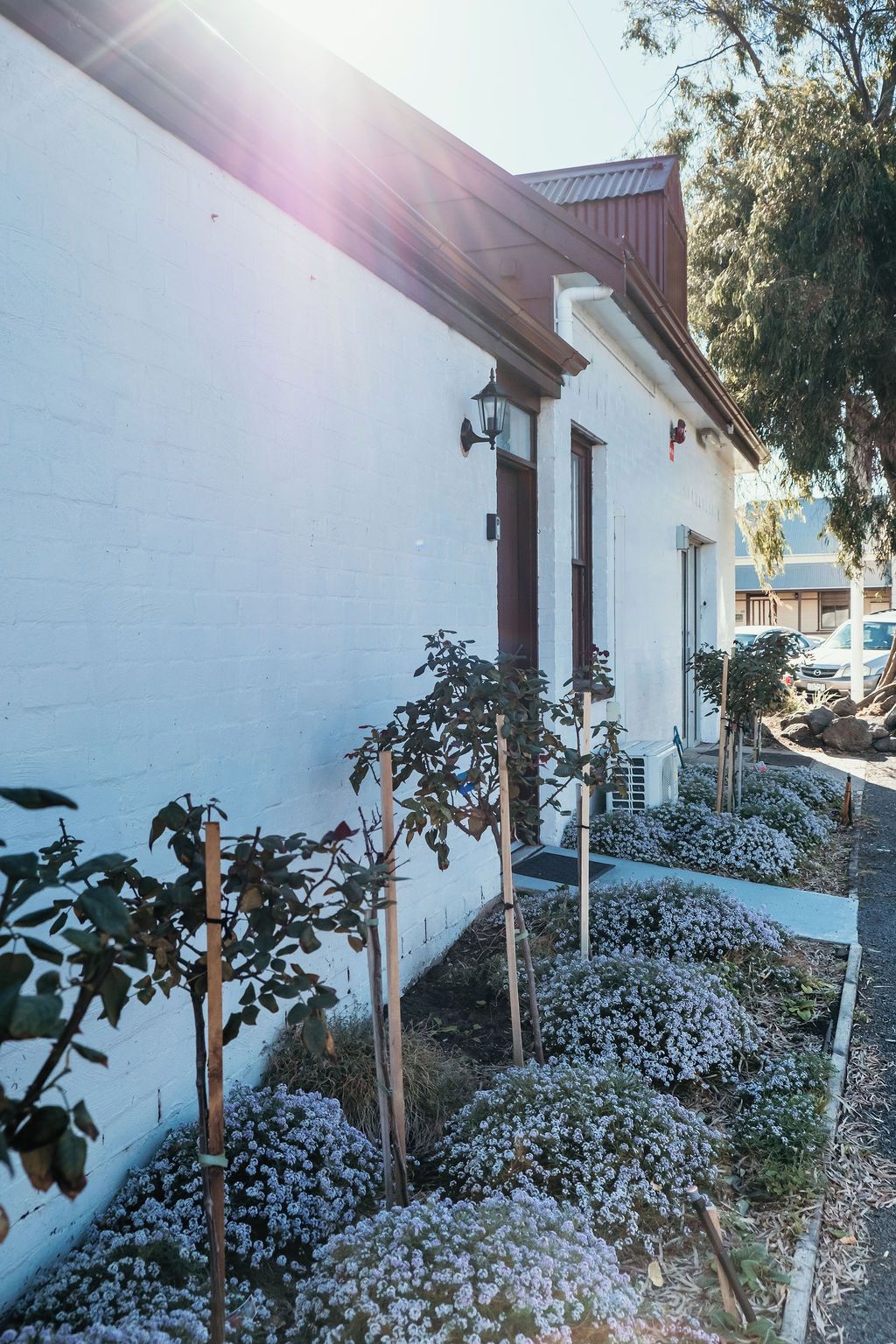 A white building with a red roof, a small garden with flowering plants, and a wooden fence.