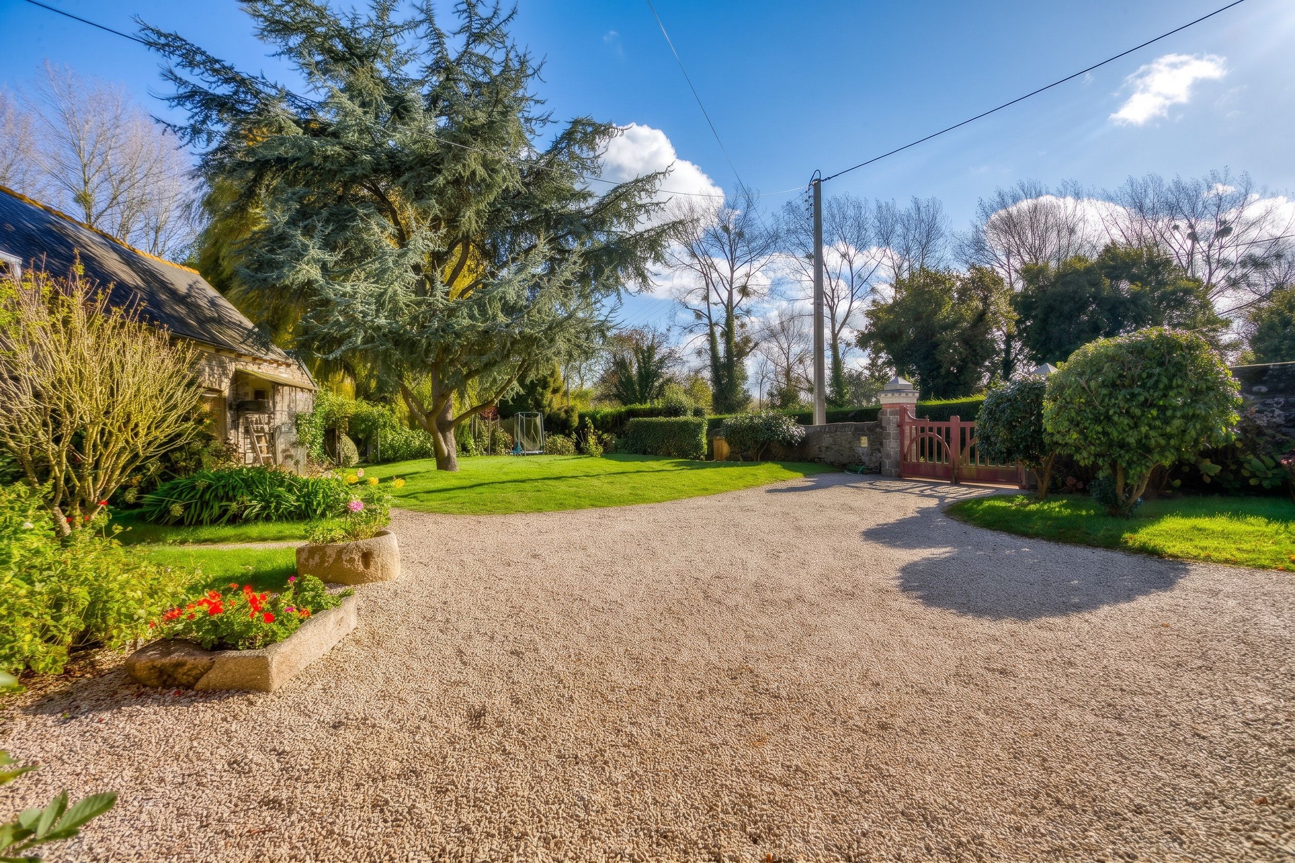 Une allée de gravier mène à une maison dotée d’un toit en pente, qui est elle-même entourée d’un jardin bien entretenu, où poussent diverses plantes et fleurs.