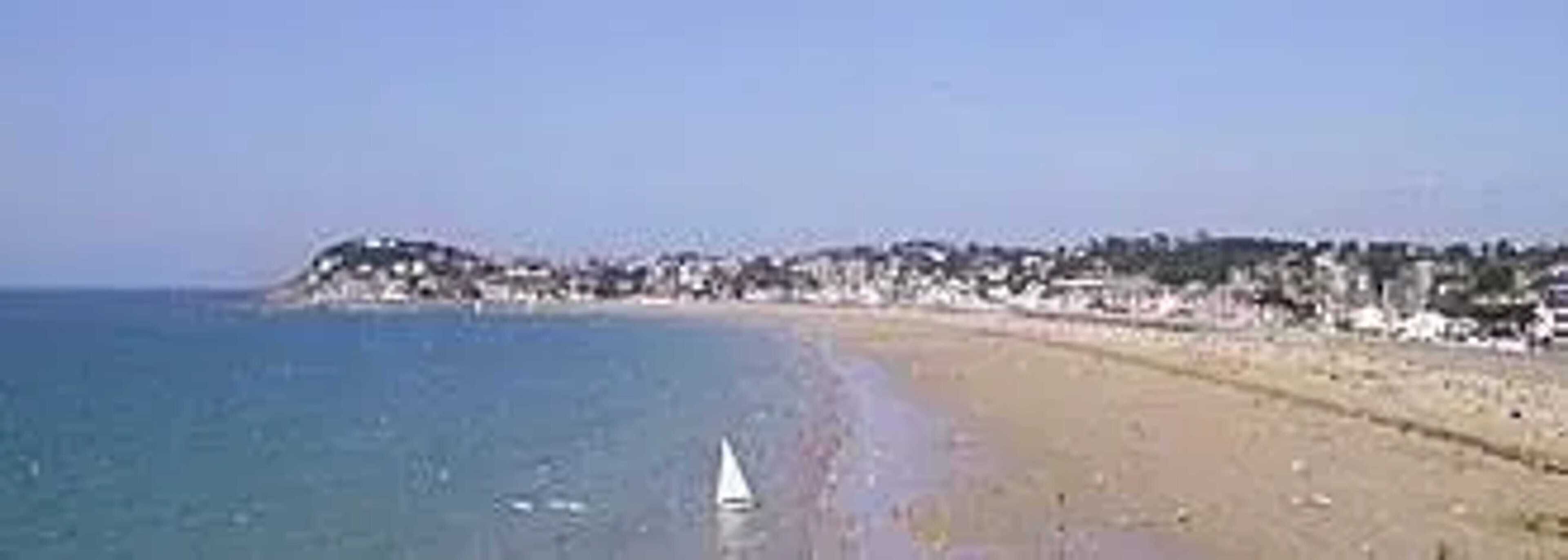 Une plage avec un voilier, un rivage sablonneux et une colline boisée en arrière-plan.