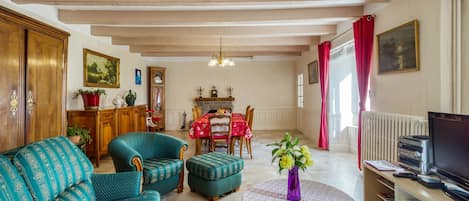 Una sala de estar con un sofá, un sillón, un reposapiés y una mesita con un jarrón. Hay una chimenea, una mesa de comedor con sillas y un radiador.