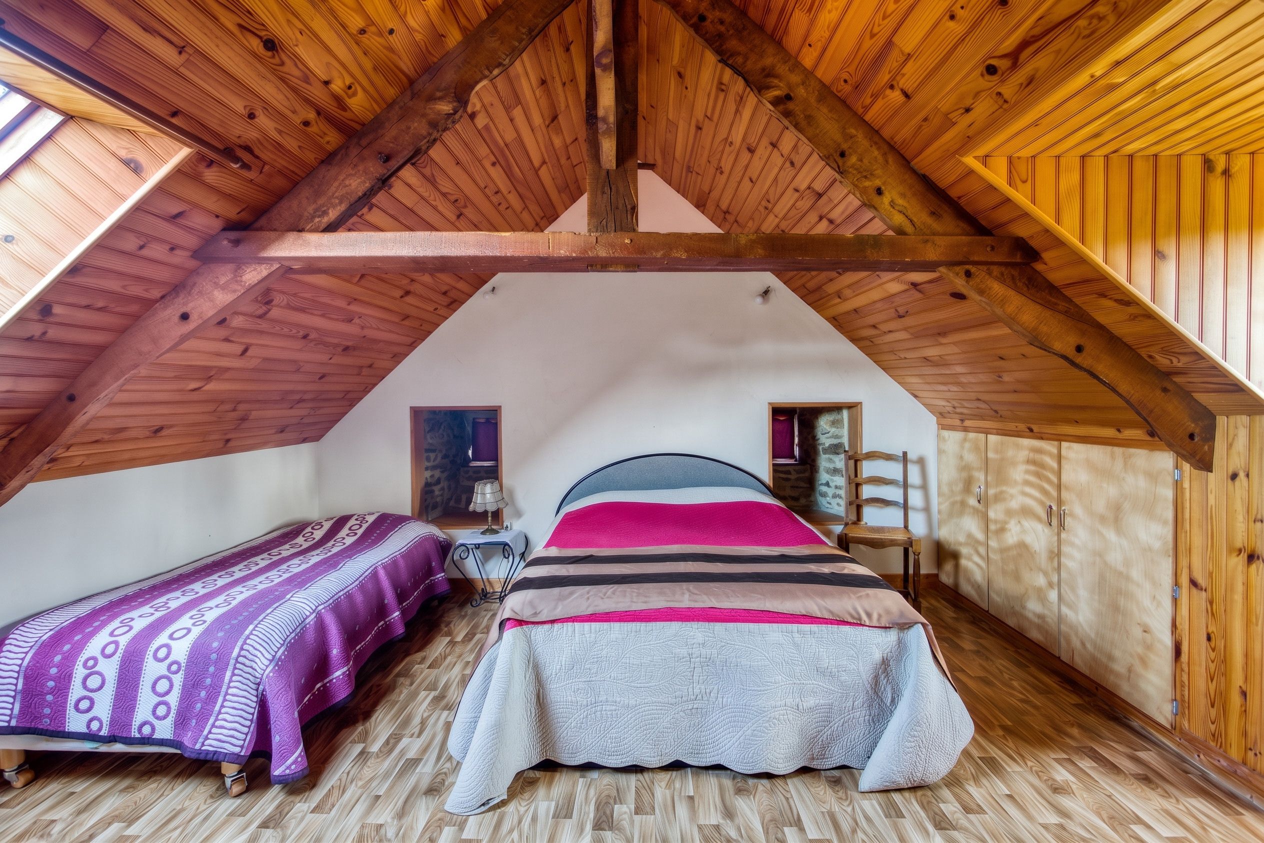 A bedroom with a sloped ceiling, wooden beams, two beds with patterned bedding, a nightstand, a lamp, and a wardrobe.