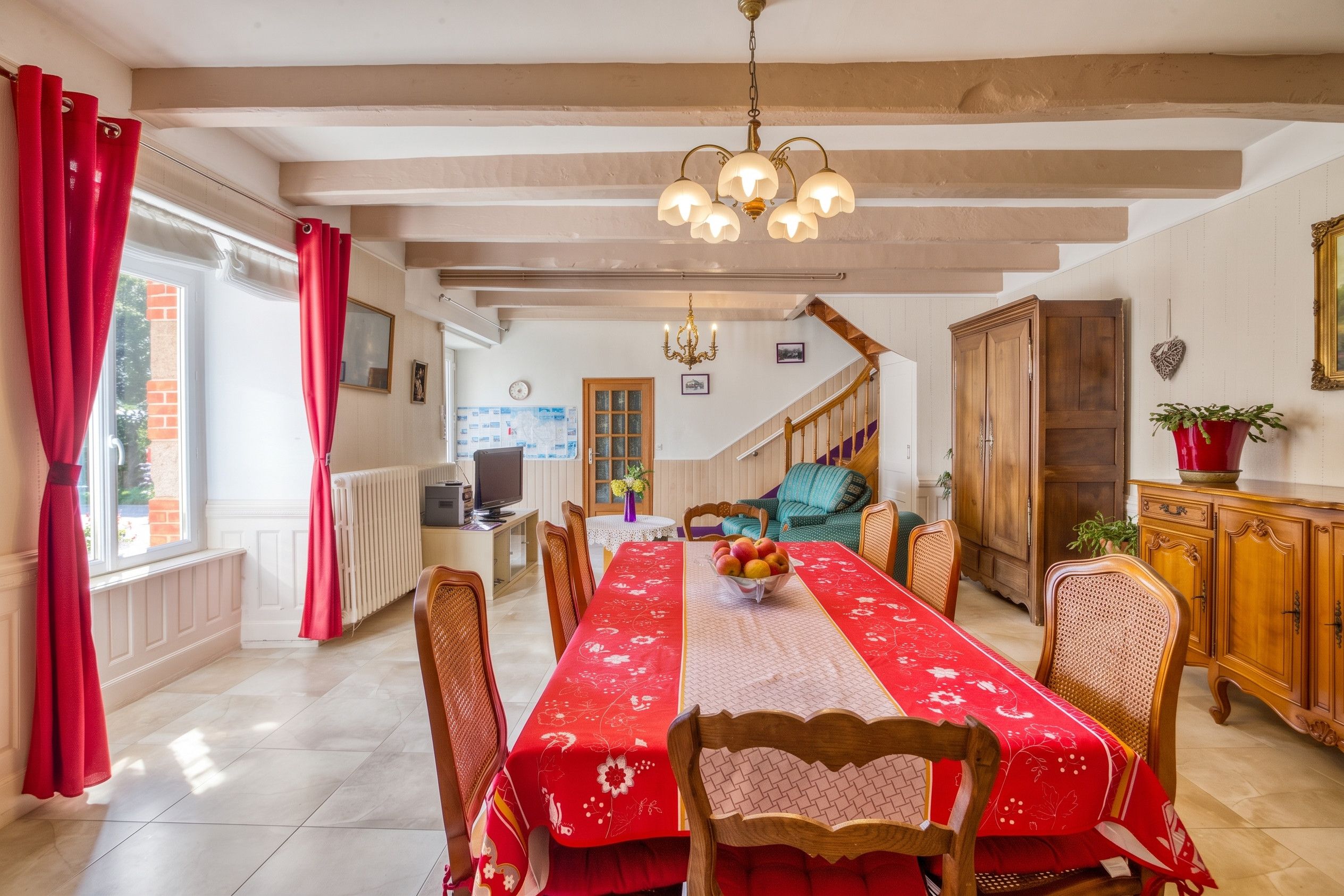 A dining area with a long table, chairs, a chandelier, and a staircase leading to another room.