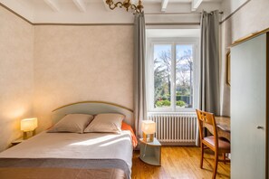 A bedroom with a bed, two bedside lamps, a radiator, a wooden chair, and a window with a view of trees. - Homerez - House in Pordic (Pordic)