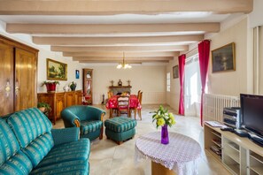 A living room with a sofa, ottoman, dining table, and chairs. There is a fireplace, a clock, and a television. - Homerez - House in Pordic (Pordic)