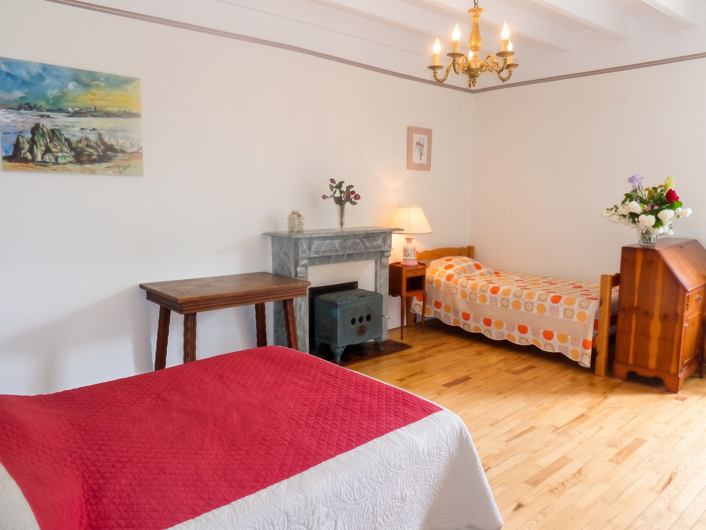 Une chambre à coucher comprenant un lit, un bureau, une chaise, une commode, une table de chevet, une lampe, un vase avec des fleurs, un tableau au mur et un lustre.