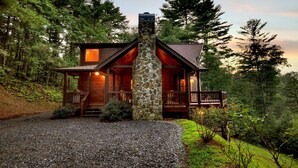 Une cabane en rondins avec une cheminée en pierre, entourée d’une forêt dense, dotée d’un porche et d’une terrasse bien visible.