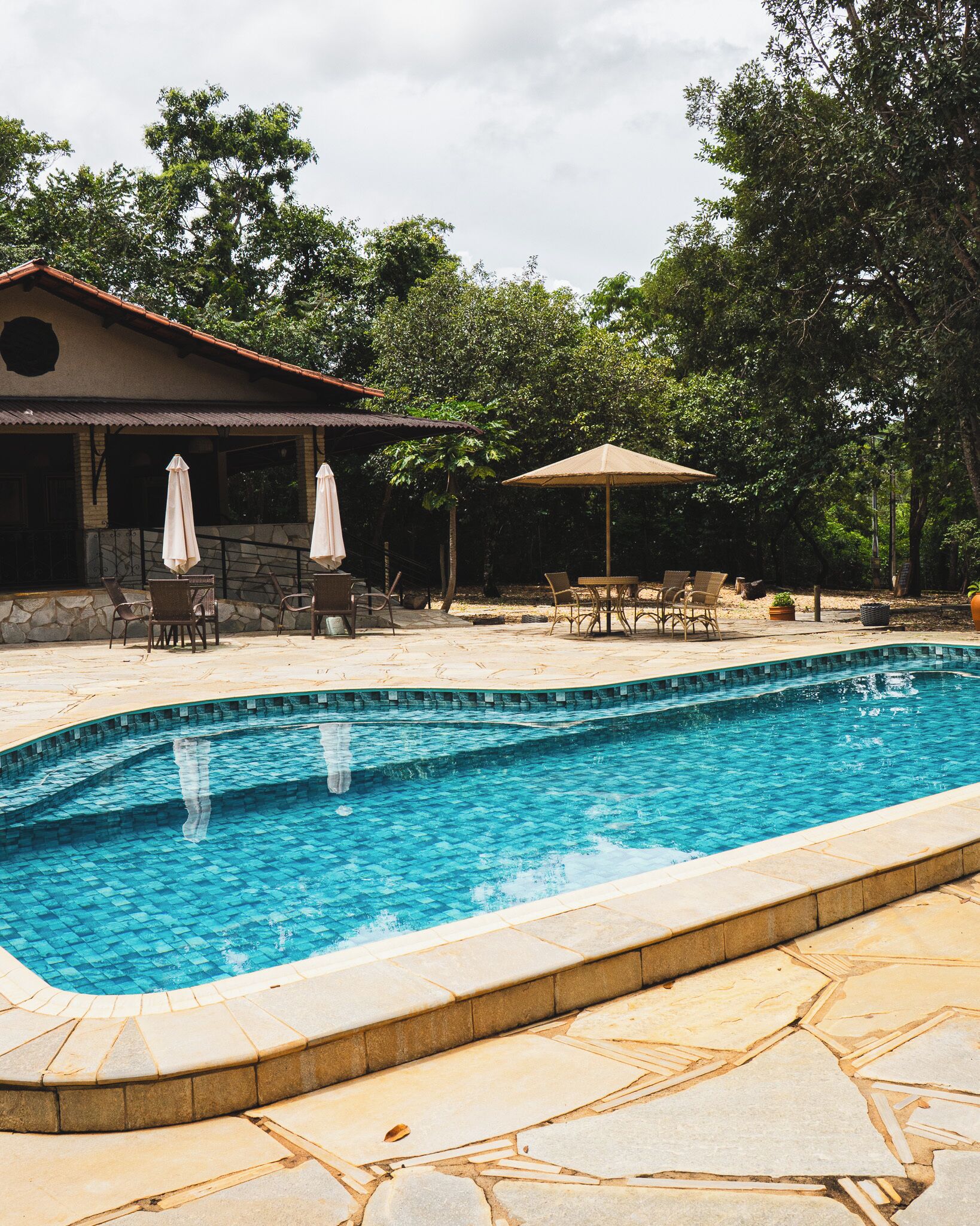 Una piscina rectangular con bordeado de azulejos, rodeada de losetas de piedra. Hay dos mesas al aire libre con sillas y sombrillas, y un edificio con tejado de azulejos al fondo.
