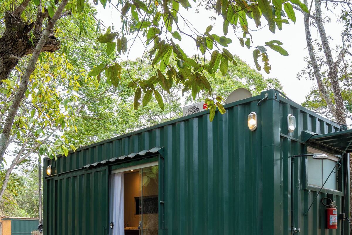 A green container-style building with a covered deck, two wicker chairs, and a fire extinguisher.