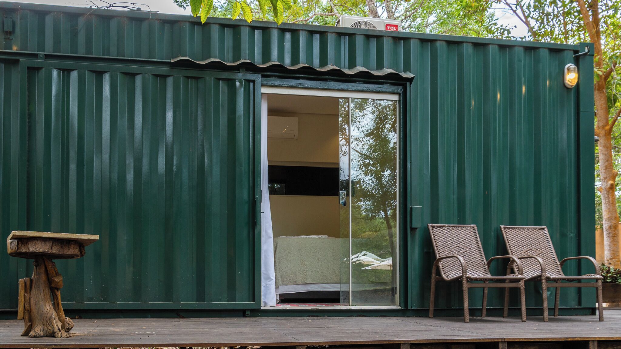 A green metal building with a sliding glass door, two wicker chairs, and a small table on a wooden deck.