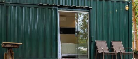 A green metal building with a sliding glass door, two wicker chairs, and a small table on a wooden deck.