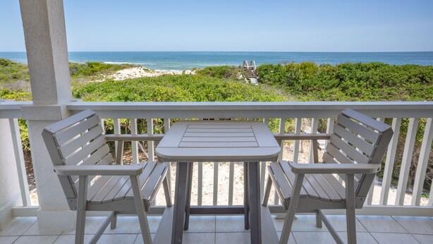 A balcony with two wooden chairs and a small table, overlooking a beach with a wooden walkway and greenery. - BEACHFRONT Beauty in the Dunes!!, 5BR/4BA, (St. George Island)
