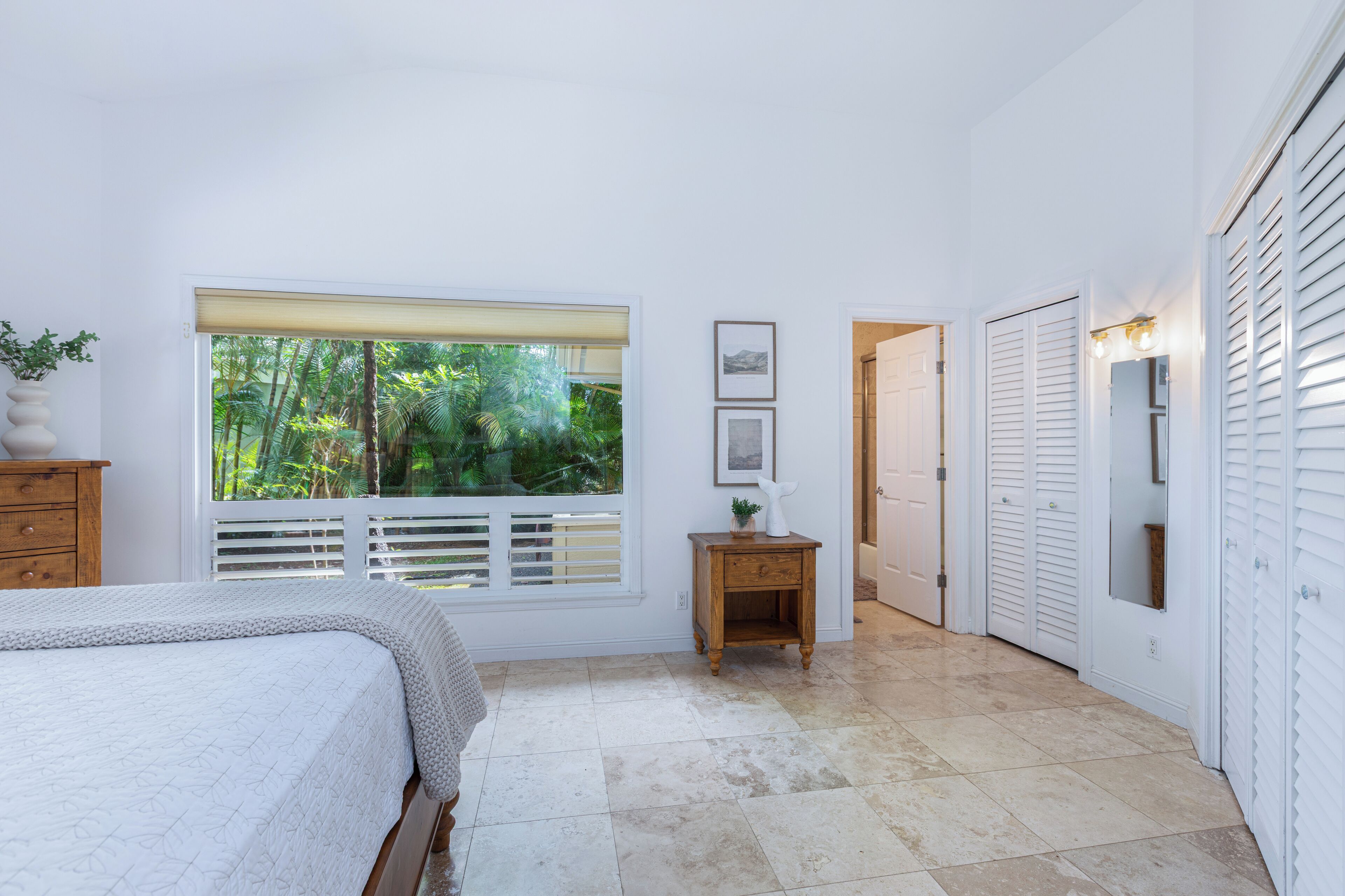 A bedroom with a large window, a bed, a nightstand, a wardrobe, and a door leading to another room.