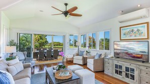 A spacious living room with a large TV, comfortable seating, and a ceiling fan. - Ohana Kamalani: Lavish Fairwayfront Home w/Pool, AC & Stunning Sunsets! (Princeville)