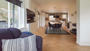 Een moderne woonkamer met een bank en een televisie, die uitkomt in een eetgedeelte met een tafel en stoelen, en een keuken met kastjes en apparatuur.