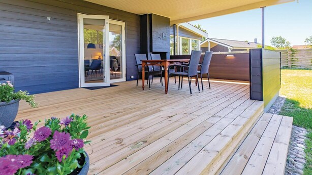 Een houten terras met een tafel en stoelen, een schuifdeur naar een binnenruimte, en een tuin met bloemen.