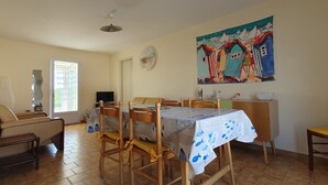 A dining area with a table set for four, surrounded by wooden chairs. A television is mounted on the wall, and a painting is visible above the table.
