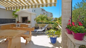 Une terrasse extérieure couverte, avec une table et des chaises en bois, des plantes en pot et une vue sur un bâtiment en arrière-plan.