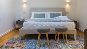 Un lit bien fait, avec une couette blanche et deux oreillers, une table de chevet ornée d’un vase et un tapis aux motifs colorés.
