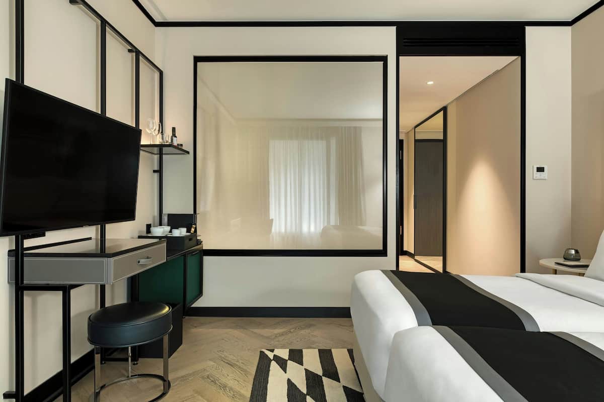 A modern hotel room with a large bed, a desk with a flat-screen TV, a small stool, and a view of the hallway and another room through a glass door.