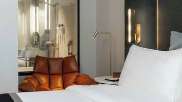 Een moderne hotelkamer met een glazen douchecabine, een ronde bijzettafel, een leren fauteuil en een wit bed.