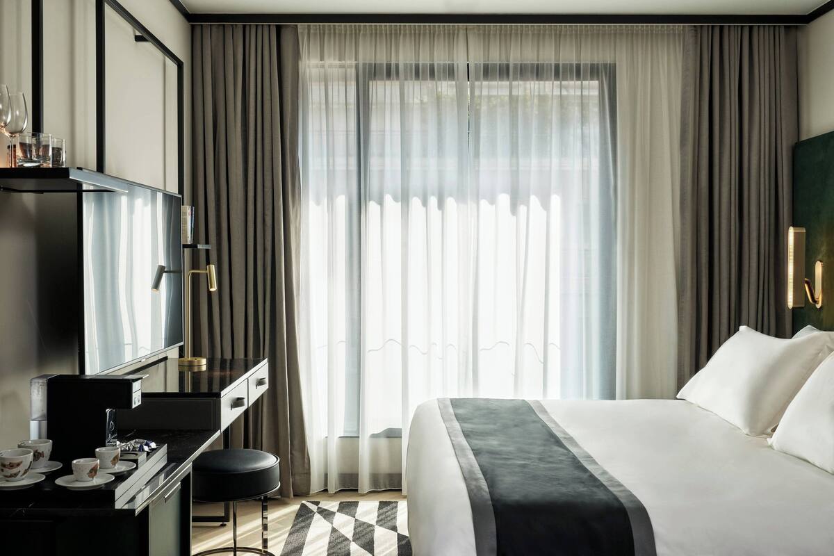 A modern hotel room with a large bed, a desk with a mirror, and a window with sheer curtains.
