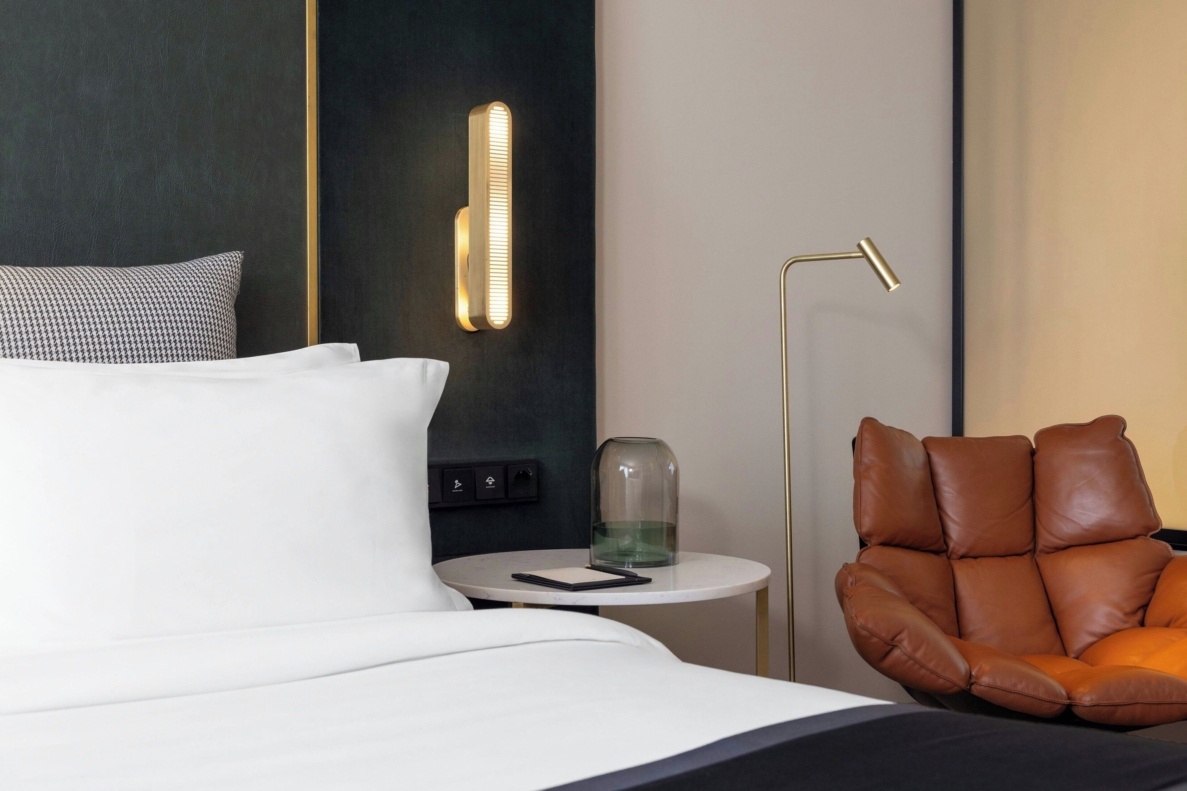 A modern hotel room with a bed, a round side table, a brown leather chair, and a wall-mounted light fixture.