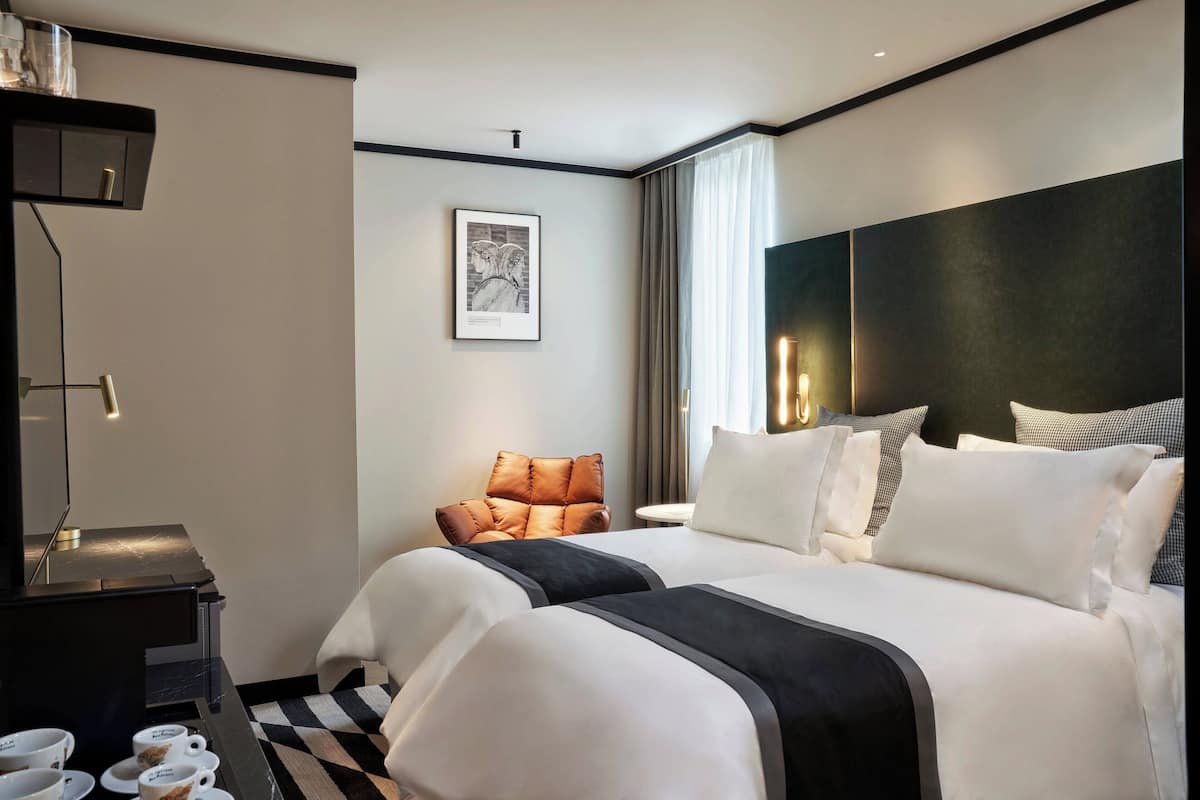 A modern hotel room with a large bed, a side table with cups, a chair, and a framed picture on the wall.