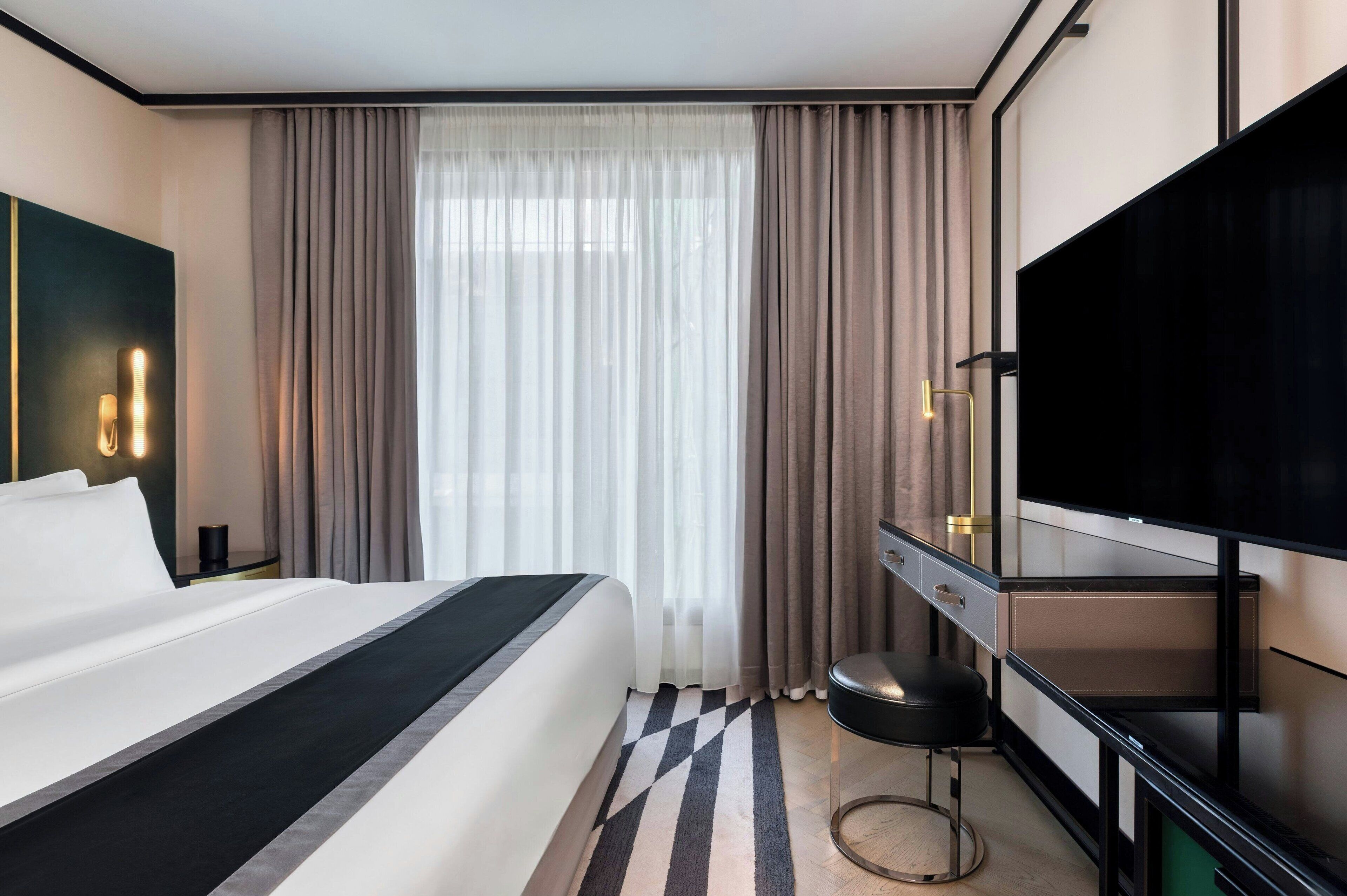 Ein modernes Hotelzimmer mit einem großen Bett, einem Schreibtisch mit Stuhl, einem Flachbildfernseher und einem großen Fenster mit Vorhängen.