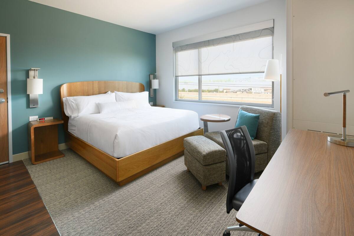 Quarto de hotel com cama, escrivaninha, cadeira, mesinha redonda, luminária e janela com vista.