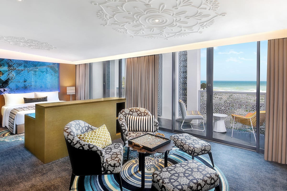 A hotel room with a bed, a desk, a chair, a small table, and a view of the ocean through large windows.