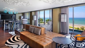 Una sala moderna con un sofá de cuero, una zona de comedor con taburetes de bar y un televisor grande. La habitación tiene vista al océano y un balcón con una silla estampada.