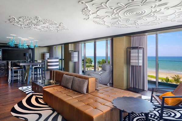 A modern living room with a leather sofa, a dining area with bar stools, and a large TV. The room has a view of the ocean and a balcony with a patterned chair.