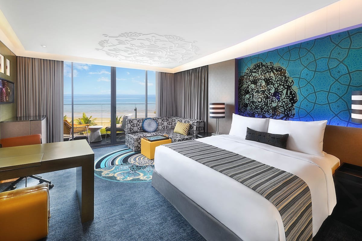 A hotel room with a large bed, a sofa, a dining table, and a view of the beach.