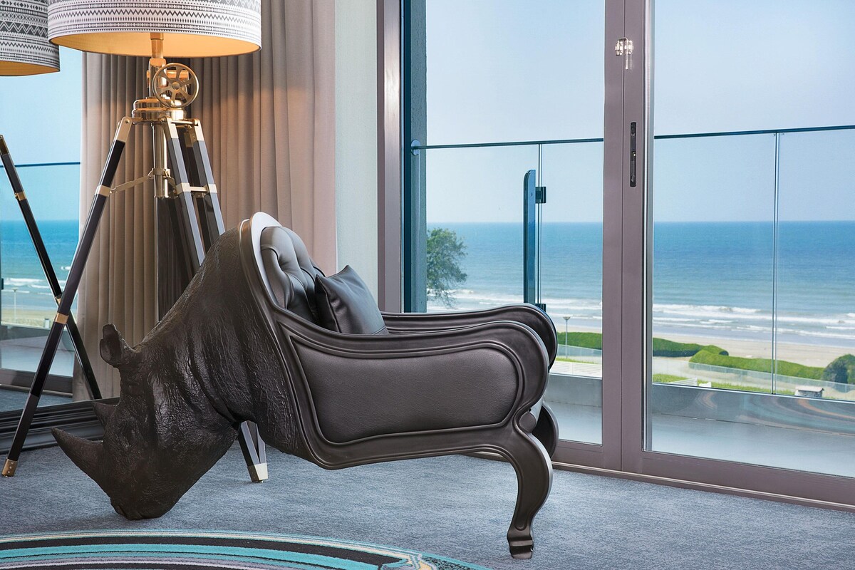 Una habitación con un amplio ventanal con vistas al océano, un sofá elegante y una lámpara con trípode.