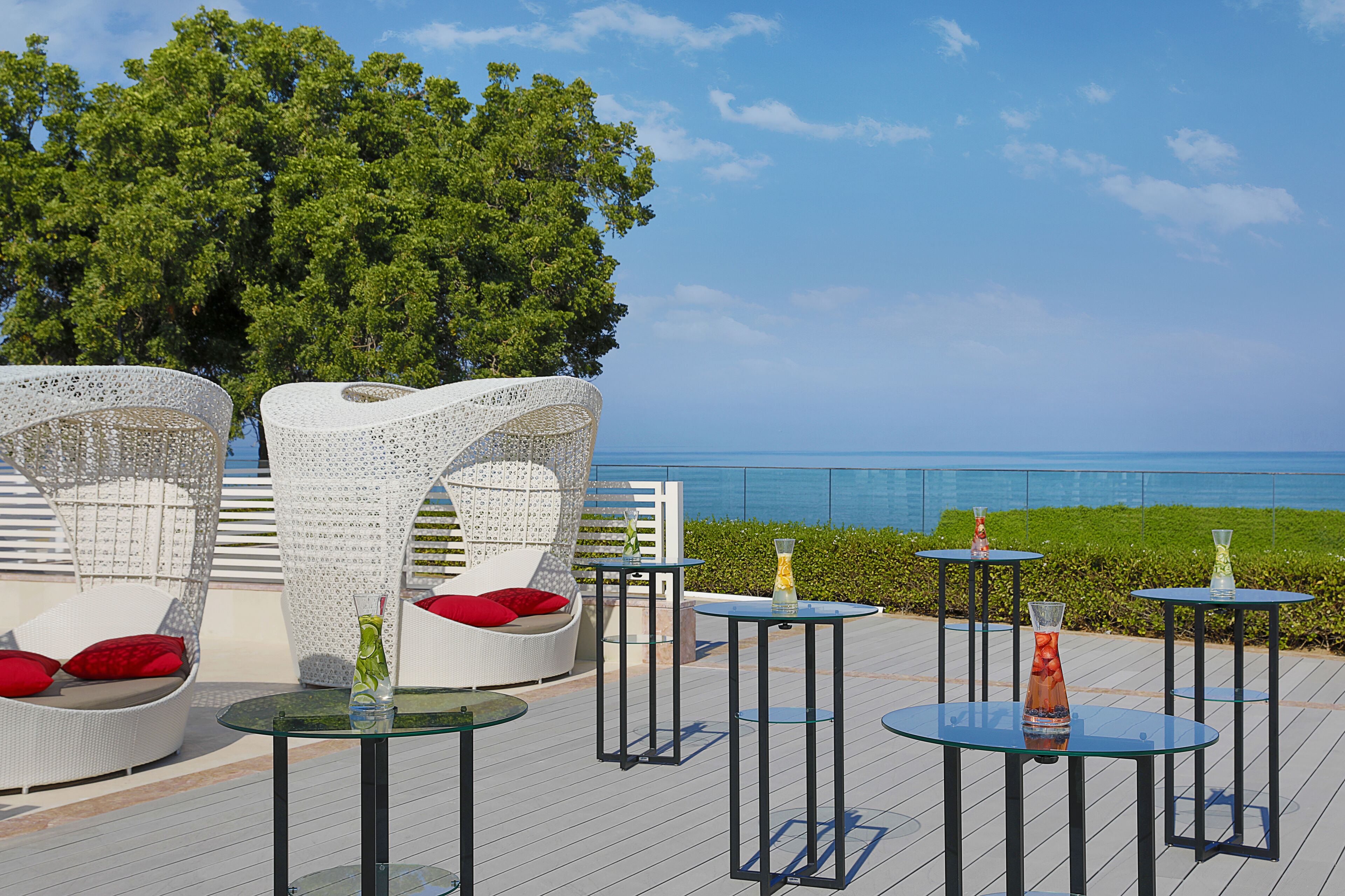 Una terraza con mobiliario de mimbre blanco, mesas de vidrio y cojines rojos.