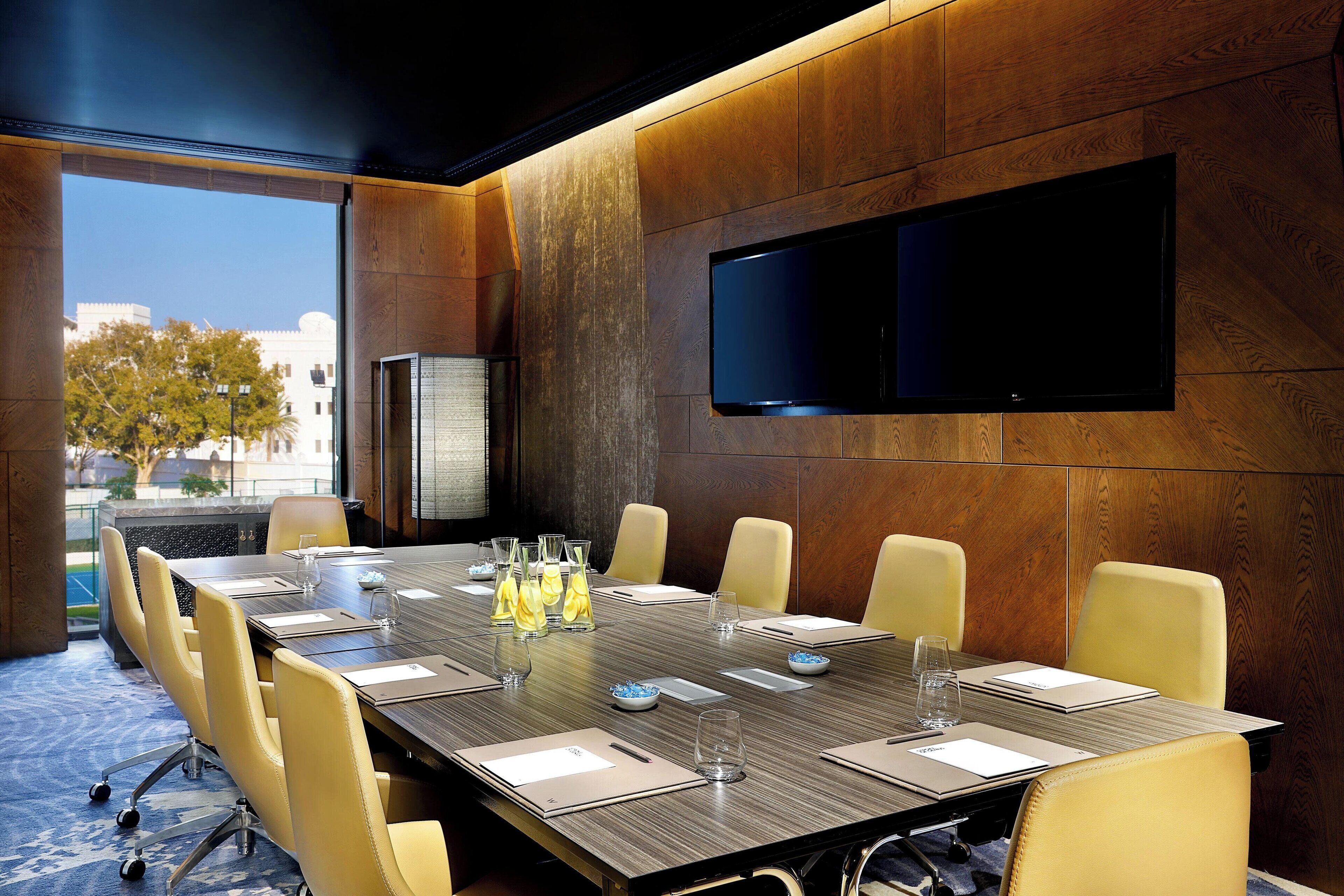 A conference room with a long table, yellow chairs, a flat-screen TV, and a view of buildings outside.