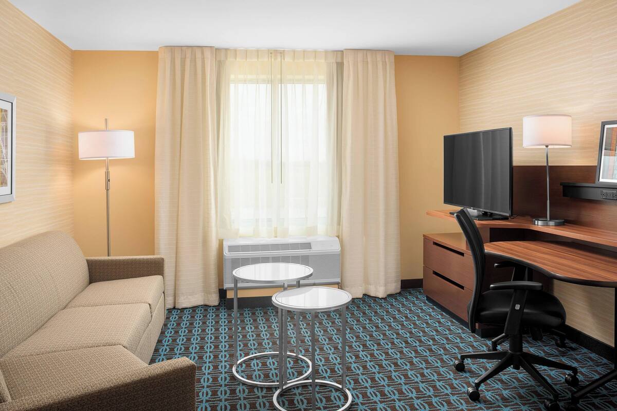 Une chambre d’hôtel avec un canapé, un bureau avec un ordinateur, deux tables de chevet, une télévision et une fenêtre avec des rideaux.