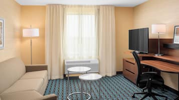 A hotel room with a sofa, a desk with a computer, two side tables, a TV, and a window with curtains.