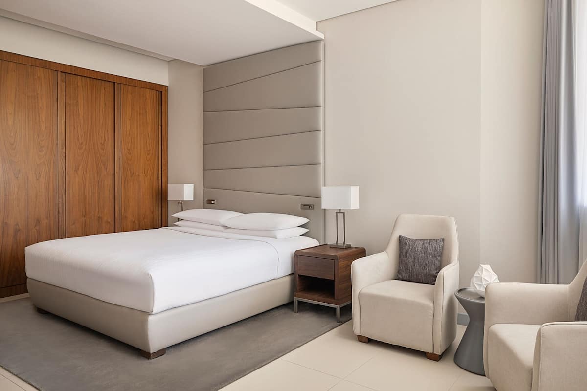 A modern hotel room with a large bed, two bedside tables, a wooden wardrobe, and a seating area with two armchairs and a side table.
