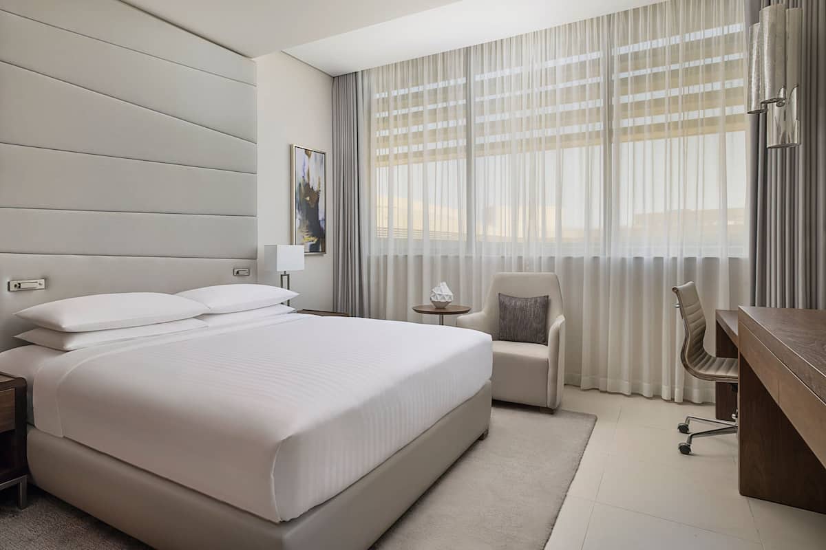 A hotel room with a large bed, a desk, a chair, and a window with sheer curtains.