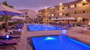 A hotel pool area with lounge chairs and umbrellas, a large pool, and a building with illuminated windows. - Marriott Riyadh Diplomatic Quarter (Riyadh)