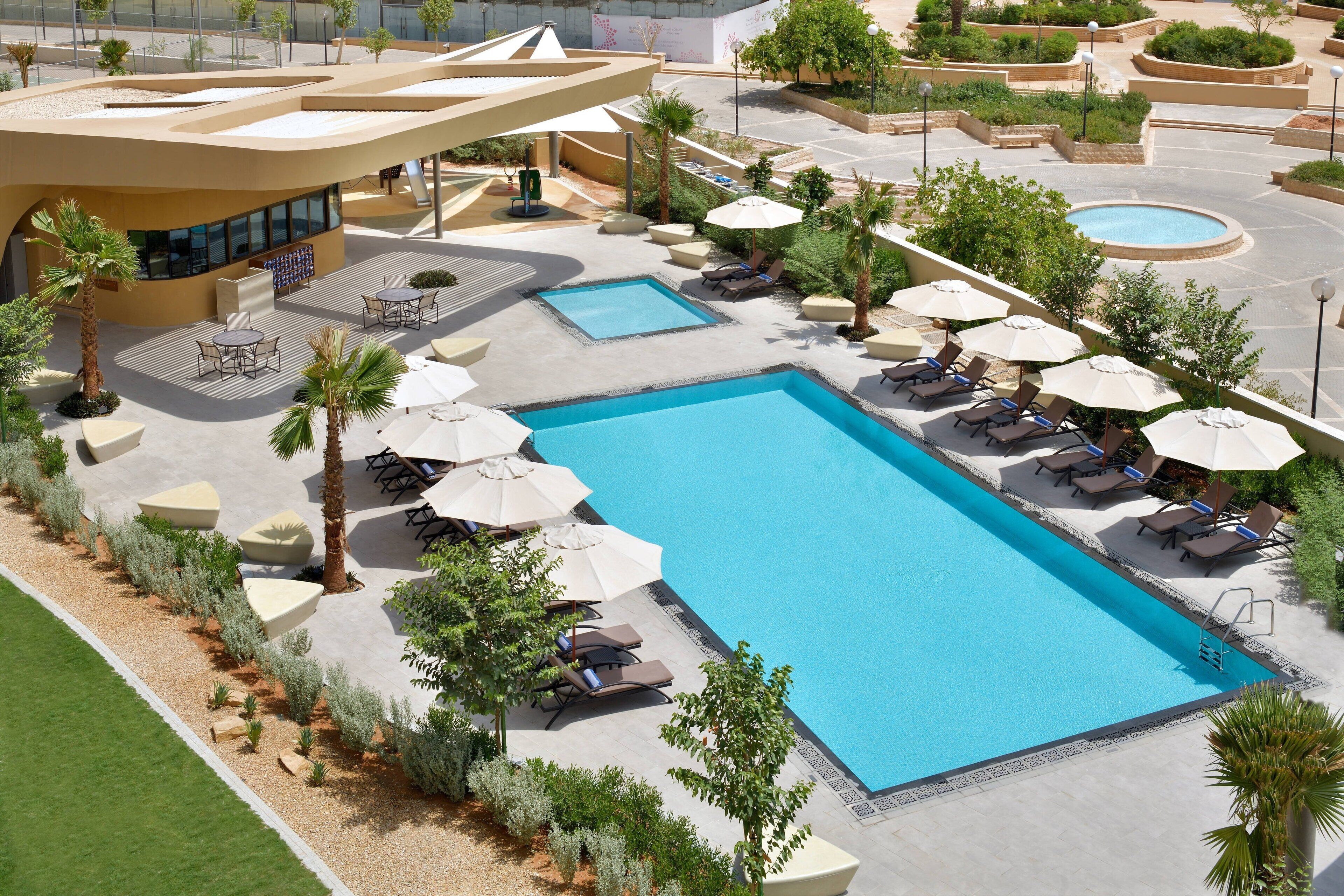 Una piscina rectangular grande con sillas de descanso y sombrillas, rodeada de palmeras y un jardín paisajístico.