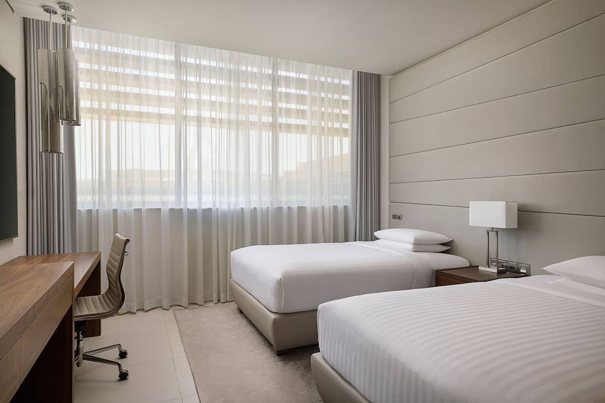 A hotel room with two beds, a desk, a chair, a TV, and a window with curtains.
