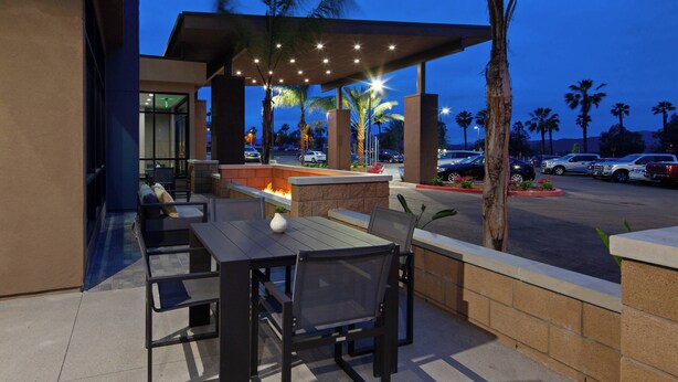 Une terrasse extérieure avec une table et des chaises, un foyer et une vue sur les voitures garées et les palmiers.