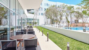 Un área de estar exterior moderna con mesas y sillas, una piscina y un edificio blanco con ventanas amplias.