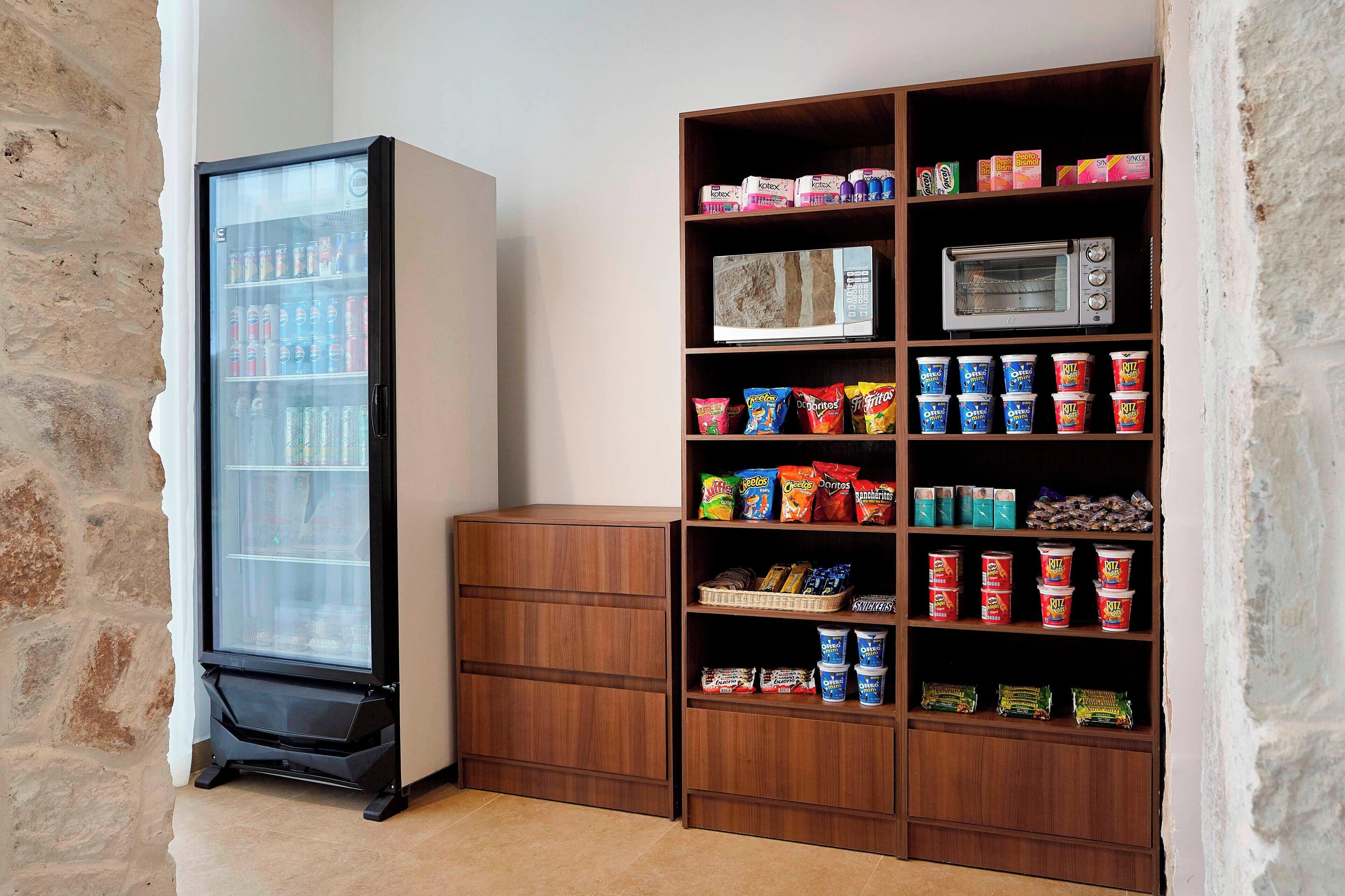 A kitchenette with a refrigerator, microwave, and shelves stocked with snacks and beverages.