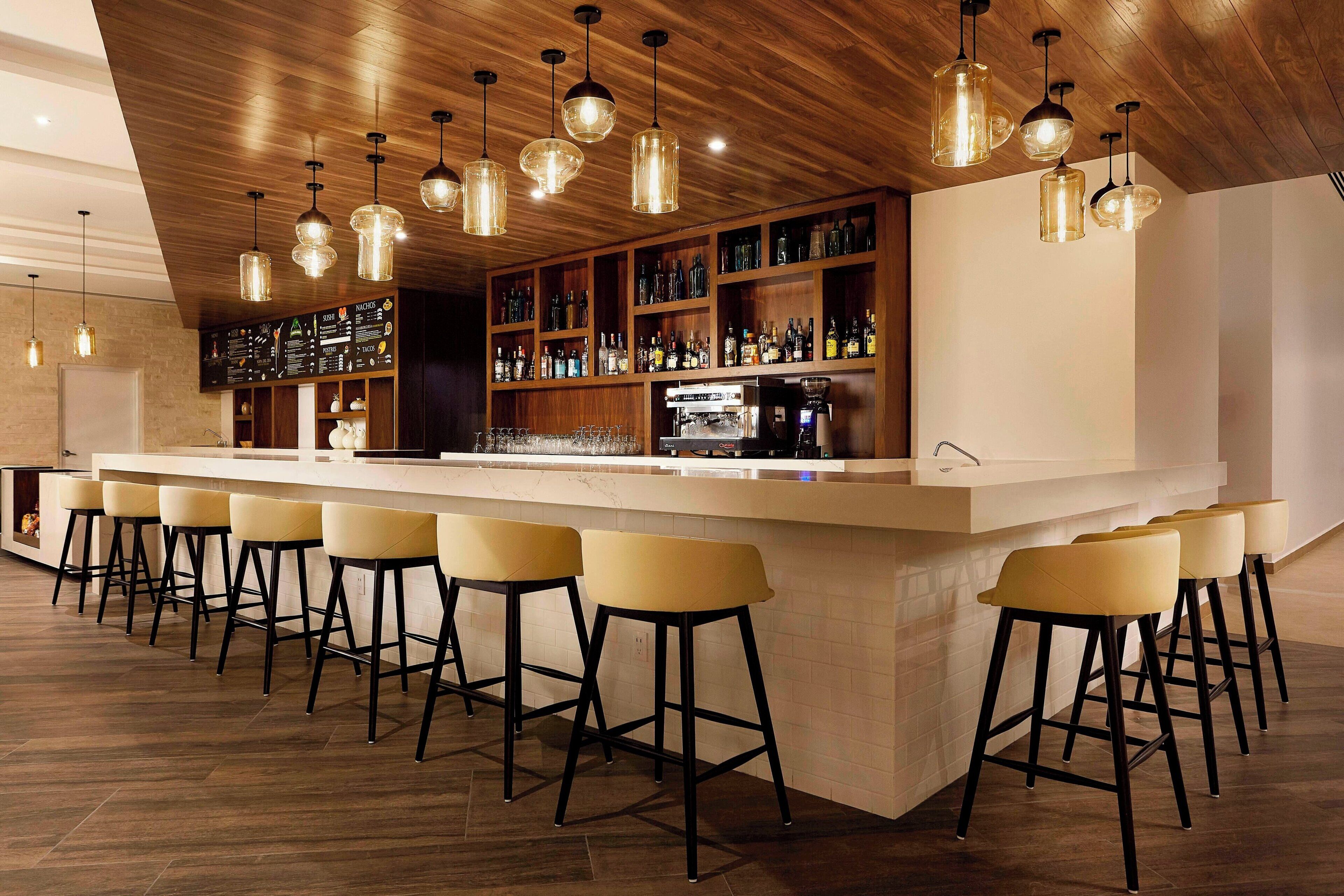 Een bar met houten plafond, hangende pendantlampen, een lange toonbank met krukken en planken vol flessen.