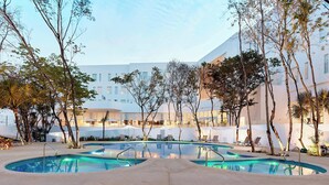 A pool area with multiple pools, lounge chairs, and umbrellas, surrounded by trees and a modern building in the background.