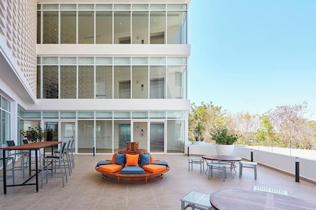 A modern outdoor seating area with a round table and chairs, a wooden table with chairs, and a large windowed wall.